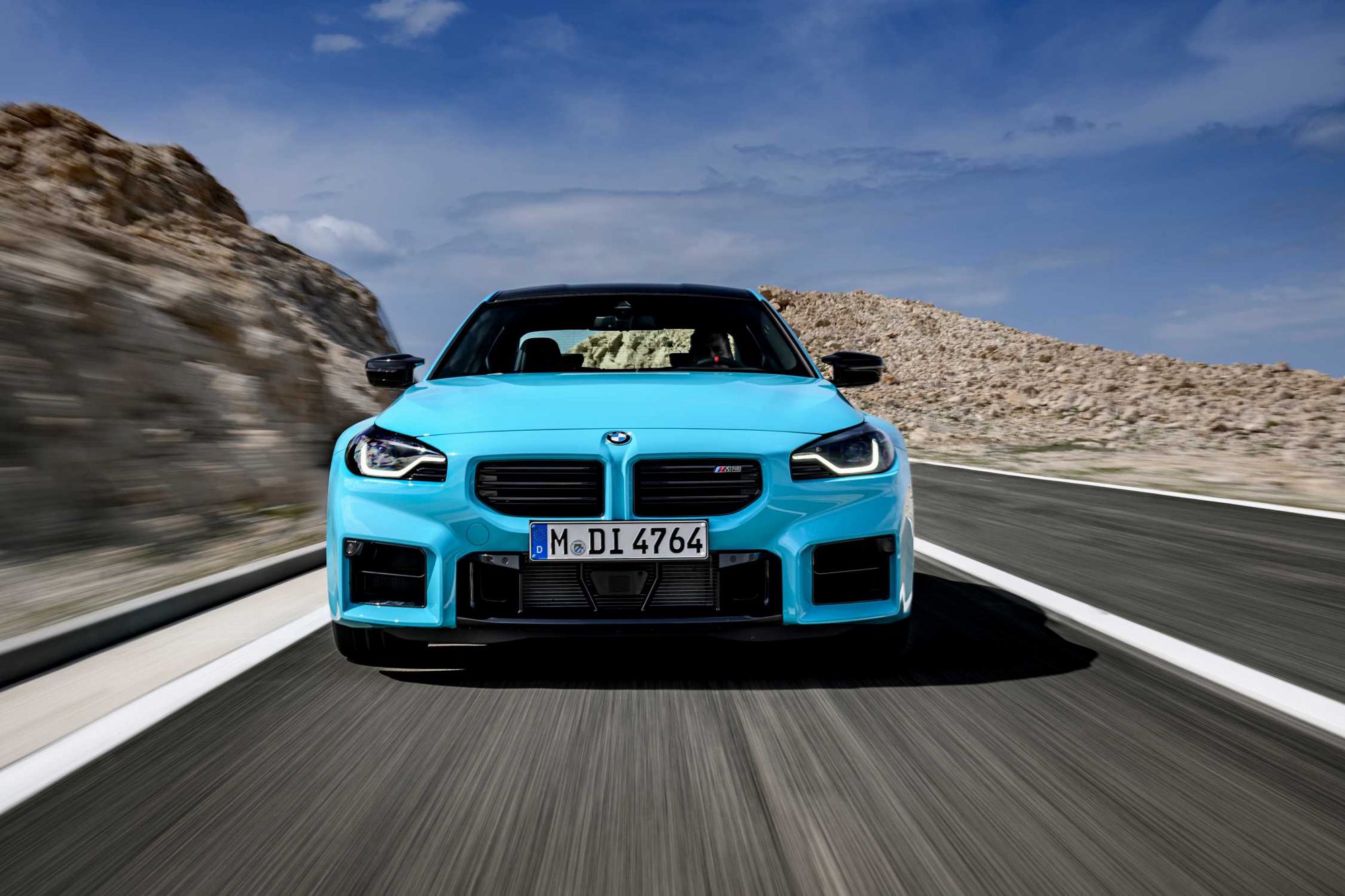 BMW M2 bleu turquoise roulant de face sur une route de campagne devant un versant rocheux clair