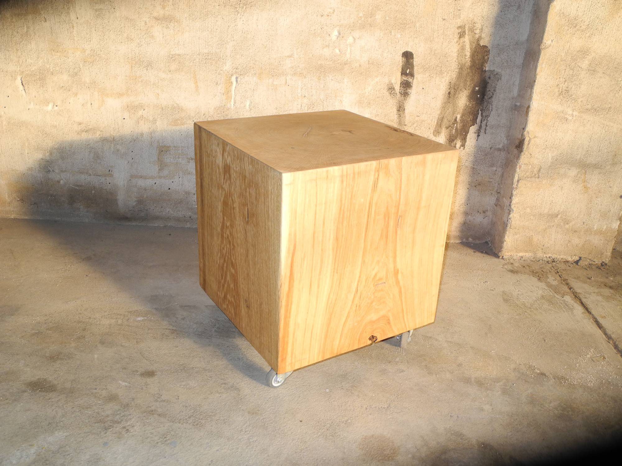 Würfelförmiger Hocker aus Massivholz mit Rollen steht schräg auf Betonboden vor einer rauen Betonwand.